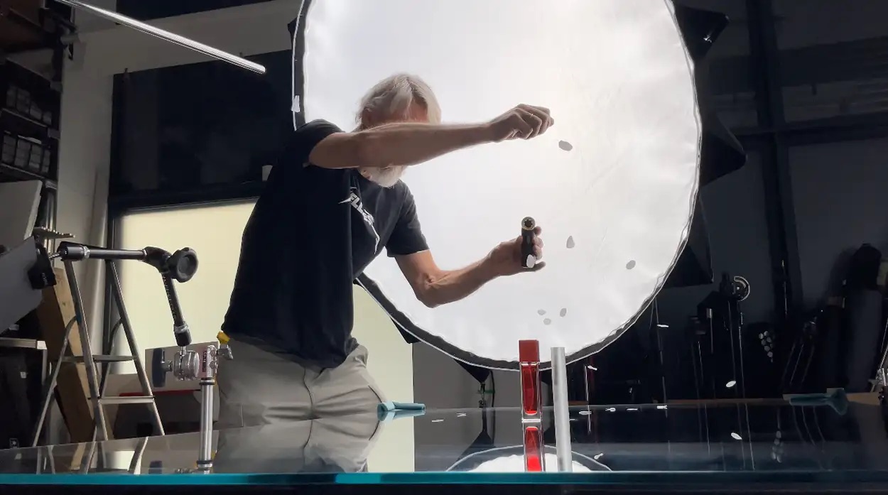 Photographe réglant un grand réflecteur circulaire devant un produit beauté rouge sur table en verre, setup packshot Greystone Studio Lyon